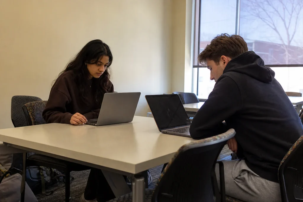Två studenter sitter mitt emot varandra vid ett bord med varsitt öppet laptop i ett universitetsbibliotek.