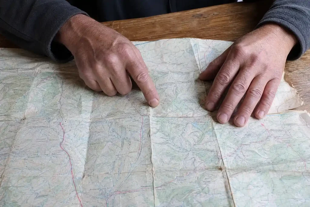 Åldrade händer som viker ut en sliten papperskarta på ett träbord och spårar en rutt med fingret.